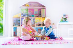 Kids playing with doll house and stuffed animal toys. Children sit on a pink rug in a play room at home or kindergarten. Toddler kid and baby with plush toy and dolls. Birthday party for little child.